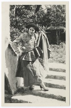 Item 0147. Young woman in matador's costume, standing on a shallow flight of stone steps. Wall 148 behind her, foliage in background. She is holding a cape in front of herself.