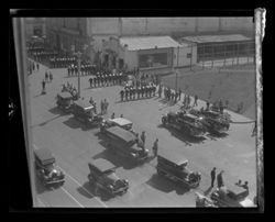 Item 0263. Either Independence Day parade or parade during athletic and military maneuvers. Lines of marching men preceding two fire trucks. Cars lined up along one side of street, people watching parade. Taken from above.
