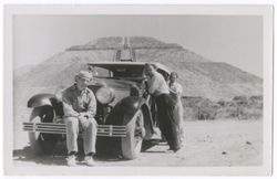 Item 39. Tissé, seated on front bumper of car with two unidentified men standing beside car. In background, the Temple of the Sun.