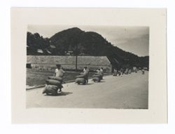 Item 0969. Line of men wheeling loaded sacks along road toward the left foreground. Long barn-like structure and mountains in background.