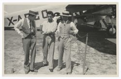 Item 0586. Three unidentified men walking toward camera. Pan-American plane in background.