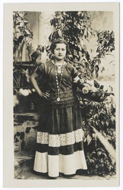 Item 04. Young woman in costume similar to those worn by young women wearing "weepeel" headdresses, holding round decorated bowl filled with flowers. Vines and large plants in pots in background, but this may be a studio shot.