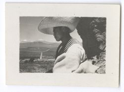 Item 0907. "Sebastian" seen in left profile, wearing sombrero, poncho, and cartridge belts across both shoulders. He is standing against a large rock on the side of a hill, with the buildings of the Hacienda visible in the far distance below.