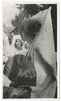 Item 0063. Profile shot of young woman in mantilla holding open fan in front of her. Other young women partially visible in background.