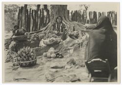 Item 0029. In center, young Indigenous woman seated among roots of large tree surrounded by round decorated baskets filled with fruit. In right foreground, a figure wrapped in a shawl and seated on a low wooden chair with back to photographer.