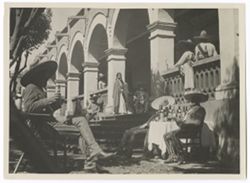 Item 0218. Scene at entrance of Hacienda. In left foreground, seated man holding a bottle (of beer?) right foreground, two men seated on either side of a table with which covering and many liquor bottles on it. Center, "Maria" (Isabel Villasenor) standing at top of steps. Three men in various attitudes behind her, and two leaning against balcony rail at upper right.