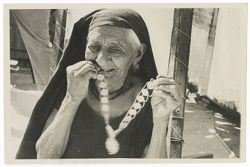 Item 0047. Close-up of an elderly Indigenous woman wearing a dark cloth veil on her head and holding up a chain and coin (?) necklace. She is smiling and biting one of the coins (?) of the necklace.
