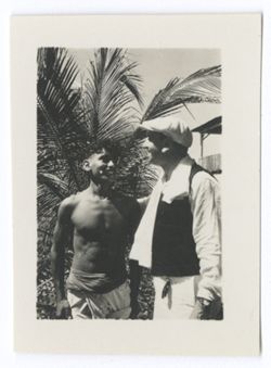 Item 1218. - 1218a. Two similar shots of young Indigenous man and unidentified man standing in front of palm tree. See also Item 478 above.