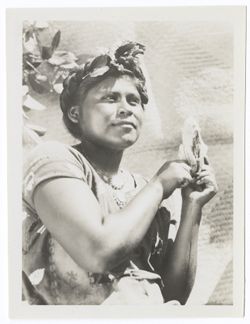 Item 0040. Young Indigenous woman holding up a half-peeled banana. Her hair is braided around her head, with a decoration of leaves and flowers over her forehead.
