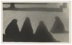 Item 0278a.  Four women, three with backs to camera, one at right in profile, all wearing dark shawls wrapped around themselves, seated in a large paved courtyard with a tree in the middle. Walls visible to the left and in the background. Closer shot - only base of tree and of rear wall visible.