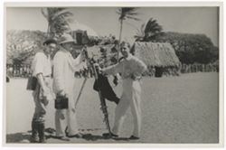 Item 0526. Dr. Best-Maugard, Kimbrough, and Tissé standing with camera on wide sandy area in front of thatched hut and other buildings.