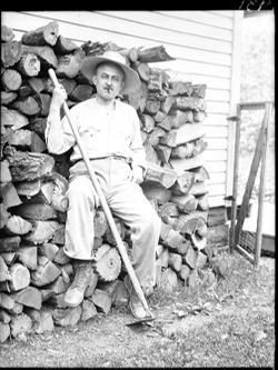 Richard Lieber with hoe on woodpile, seated