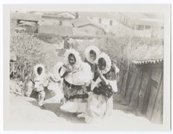 Five young Indigenous women wearing "sunflower" headdresses climbing a hill. Village in background.
