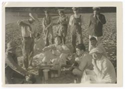 Item 0513. Eisenstein seated, center, amid a group of 10 men on a beach, some seated, some standing. They are eating and drinking from ceramic cups.