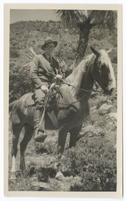 Item 0576. Middle-aged man wearing business suit, fedora, and glasses on horseback. Rocky hillside with cactus and low brush in background.