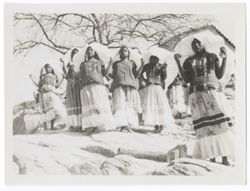 Item 0022. Group of eight Indigenous women in “weepeels” standing on rocks. Leafless tree and building in background.