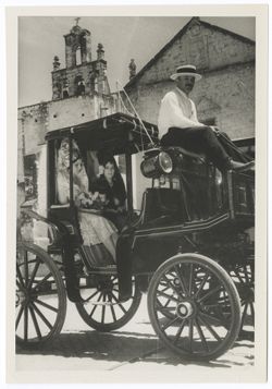 Item 0073. Two young women seated in a coach: driver holding reins in the box. One woman is dressed in white, with a white mantilla and holds flowers; the other is in a dark mantilla and dress. Church in background.