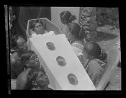 Item 0186a. Mayan funeral sequence. Two similar views of three men (left) and three women seated on either side of the coffin. Three bowls of grain (?) in a row on top of the coffin, head of "corpse" visible. Head and shoulders of "corpse" visible. In upper corner, hand holding one side of a large cloth (?) which is hung as background.