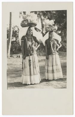 Item 02. Two young women in "weepeel" headdresses standing in open area with round, decorated bowls containing small lacy flags on their heads. Same location, but not same women, as in photo Item 28.