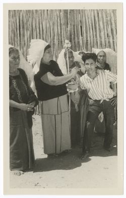 Item 0043. Group of five older Indigenous women in "weepeel" headdresses. The woman second from left is applying paint or makeup to the face of a half-crouching young Indigenous man. The woman to her left holds a staff with flowers on the end.