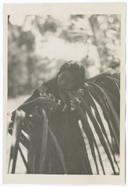 Item 0038. Young Indigenous woman leaning head against palm branch, a garland of flowers in her hand.