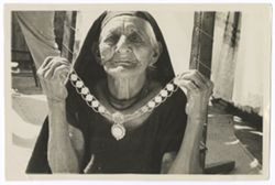 Item 0046. Close-up of an elderly Indigenous woman wearing a dark cloth veil on her head and holding up a chain and coin (?) necklace. Her eyes are open and she has a more serious expression.