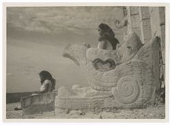 Item 0167. Stone serpent's head at entrance to upper temple, Temple of the Warriors. An Indigenous woman stands on the far side of it, facing left. In front of it, another Indigenous woman is seated on a stone slab, she also faces left.