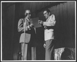 Hoagy Carmichael on stage receiving a document from an unidentified man.