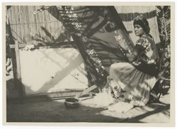 Item 0042. Young Indigenous woman seated, right, in front of a large wooden frame on which is stretched a piece of cloth, she appears to be sewing or embroidering on it. The pattern is similar to that on the blouse which she is wearing. To her left is a smaller frame with another piece of cloth on it. In the background is a fence similar to the one seen in Items 35-36 above.
