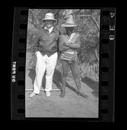 Item 0567. Three men grouped around a camera. Village with thatched roofs, palm trees in background.