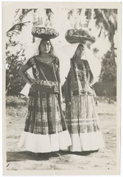 Item 0028. Full-length shot of two young Indigenous women. Both wear "weepeels" and carry round decorated baskets containing small lacy flags on their heads. Tropical foliage in background. These women are seen individually in Items 21 and 33.