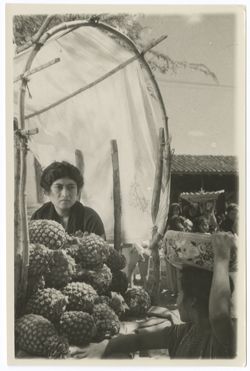 Item 0041. Indigenous woman seated beneath a stick framework over part of which a cloth has been draped as a sunshade. On a counter in front of her is a large pile of pineapples. In the lower right corner, another Indigenous woman is standing with her right arm on the counter and her left hand steadying a round decorated bowl on her head. In the background, Indigenous women and children and a low building with a tile roof.
