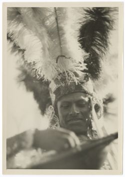 Head and shoulders of Indian in headdress with beaded headband and pendants and upright feather crown. He is playing a guitar.