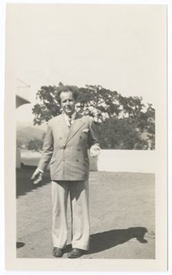 Item 0303. Full-length photo of Eisenstein standing on a lawn or terrace, gesturing with both hands. Corner of house with balcony just visible at upper left, trees and hills in background.