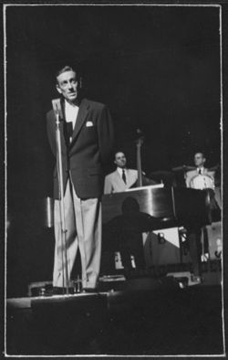 Hoagy Carmichael speaking into a microphone on stage with two unidentified musicians in the background.