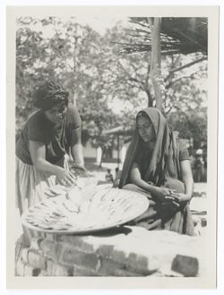Two Indigenous women, one at left putting food onto a large round tray, one at right seated watching her. Low brick wall in foreground, trees in background.