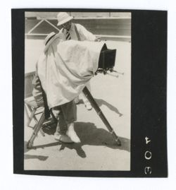 Item 0981. Unidentified man seated at camera, with cloth covering most of camera and him. Eisenstein standing in background.