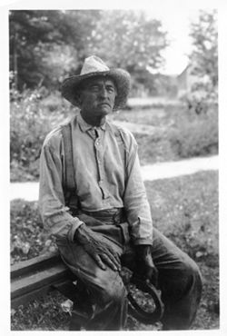 Seated man in straw hat, holding pair of horseshoes