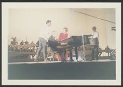 Hoagy Carmichael on stage playing the piano, with unidentified people standing nearby, and an orchestra in the background.