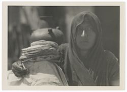Item 0192. Indigenous woman, head and shoulders wrapped in a dark cloth shawl, with her right arm around a cloth-draped object on top of which rests a pile of tortillas. In the center background is a pottery jug with three small round handles.