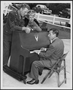 Hoagy Carmichael playing the piano on a tennis court, with an unidentified soldier and woman looking on.