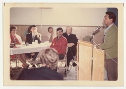 Cesar Chavez at lectern addressing people at tables