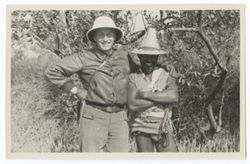 Item 0322. Alexandrov, left, with arm around shoulders of Indigenous person wearing high-peaked straw hat. Foliage in background.