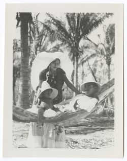 Item 0008. Girl in “weepeel” headdress leaning over man lying in hammock with baby in his lap. Tropical foliage in background. On Back, written in blue ink: “Our leading lady in Tehuantepec. None of these are for publication. Others for that shortly.” Directly below, written in pencil: “[H.S. Kimbrough writing].”