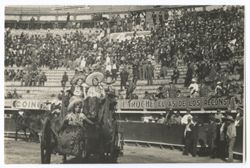 Item 0144. Three young women in sombreros and folk costumes riding in the back of a horse-drawn coach in a bullring. Spectators in the stands appear to be mostly men in business suits, unlike the mixed, casual crowds seen in the preceding photos.