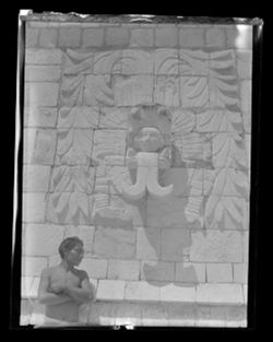 Item 0171b. Various similar views of young Indigenous man posing against a wall of the upper temple, Temple of the Warriors, and of the stone head above him on the wall. Young man from waist up, his head turned to the right.