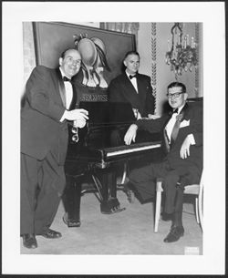 Three unidentified men leaning on piano.