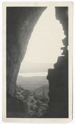Item 0282. View form cleft in hillside or entrance to the cave, looking out to a lake in the valley below. Various types of cactus on hillside.