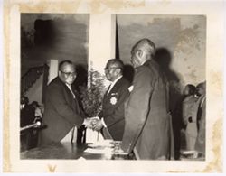 President William V. S. Tubman shakes hand with man across desk with Richard Henries and others in background