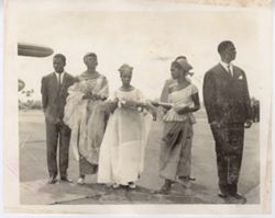 Women in dresses walk on runway holding flowers and large wooden spoon, escorted by men in suits